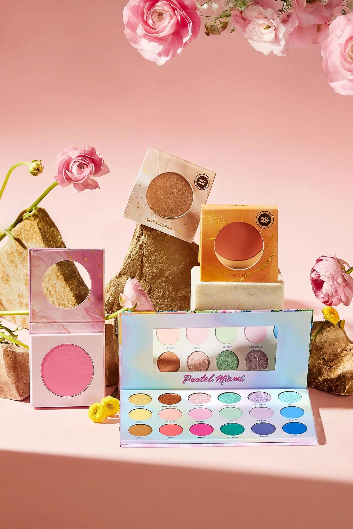 Lidschattenpalette Pastel Miami, Rouge und einzelne Mexmo-Lidschatten auf Steinen zwischen Ranunkeln vor rosa Hintergrund, Kosmetik-Werbeshooting